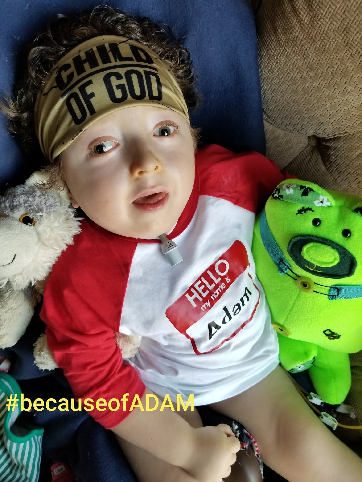Adam smiling in a "Child of God" headband and a "Hello, my name is Adam" shirt, surrounded by his stuffed animals